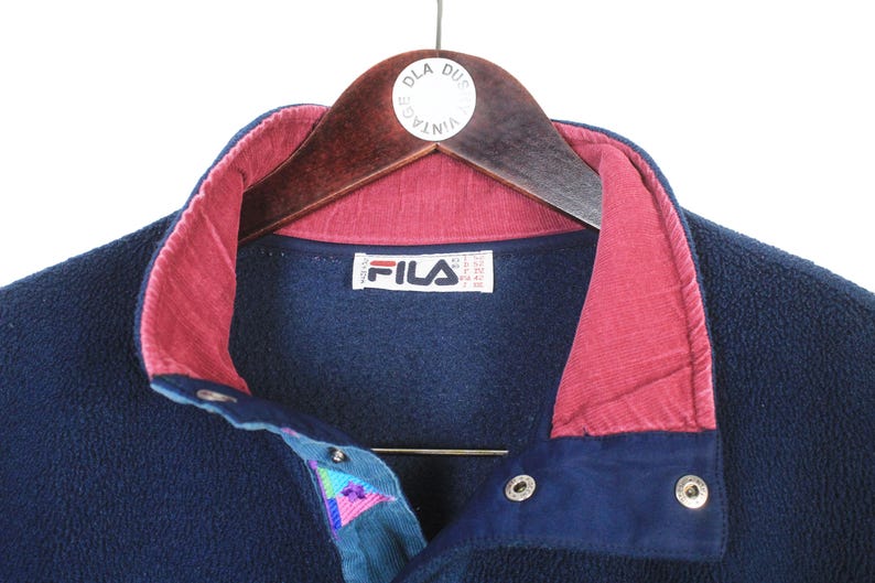 K&ouml;nnte beinhalten: Marineblaue Fleecejacke mit bordeauxfarbenem Kragen und Akzenten. Die Jacke hat einen Knopfverschluss und ein teal- und lilafarbenes Musterdetail. Das Fila-Logo ist auf dem Etikett sichtbar. Die Jacke h&auml;ngt an einem Holzkleiderb&uuml;gel.