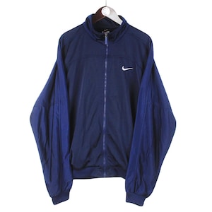 May include: Navy blue Nike track jacket with a white Nike swoosh logo on the chest. The jacket has a full zipper, a stand-up collar, and dark blue sleeves. The jacket is hanging on a white hanger against a white background.