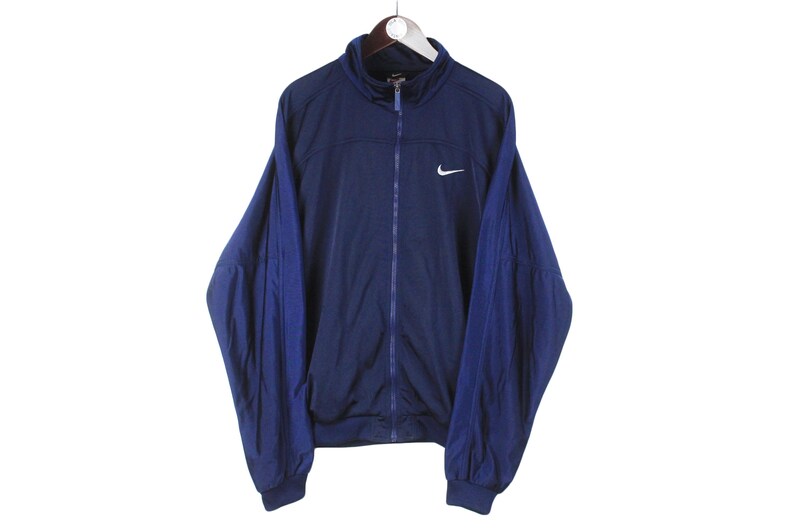 May include: Navy blue Nike track jacket with a white Nike swoosh logo on the chest. The jacket has a full zipper, a stand-up collar, and dark blue sleeves. The jacket is hanging on a white hanger against a white background.
