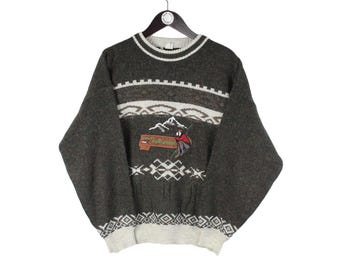 May include: A charcoal gray long-sleeve sweater featuring a white ribbed crew neckline and hem. The chest displays a graphic of a skier on a mountain with the text "Skiing is believing" on a red banner, surrounded by white geometric patterns.