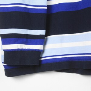 Puede incluir: Primer plano de una camisa de manga larga con rayas horizontales en tonos azules y blancos. El pu&ntilde;o es de color azul marino liso. La tela de la camisa parece ser un material de punto suave.