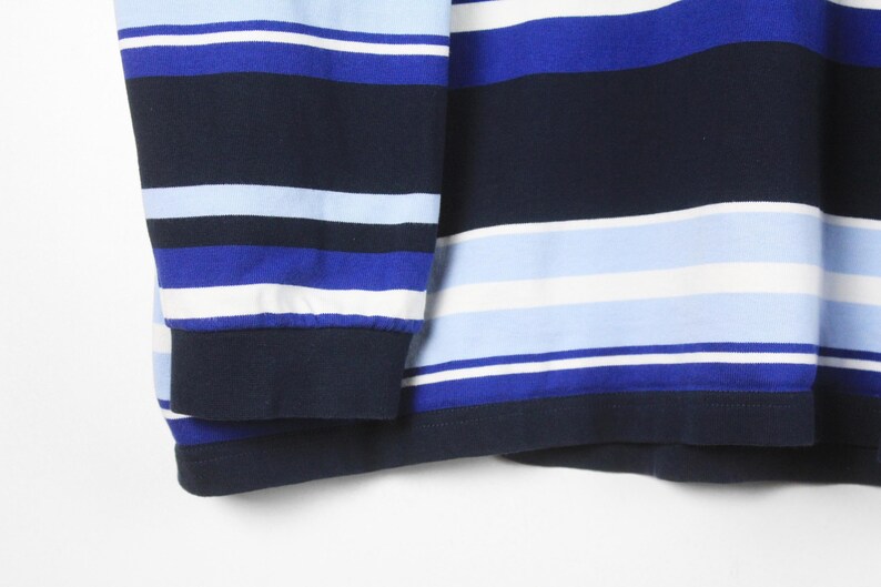 Puede incluir: Primer plano de una camisa de manga larga con rayas horizontales en tonos azules y blancos. El pu&ntilde;o es de color azul marino liso. La tela de la camisa parece ser un material de punto suave.