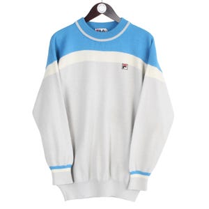 Puede incluir: Un su&eacute;ter Fila vintage en azul claro, blanco y gris, colgado de una percha de madera. El su&eacute;ter presenta un dise&ntilde;o de bloques de color con el logotipo de Fila en el pecho. Los pu&ntilde;os y el cuello tienen rayas azules y blancas.