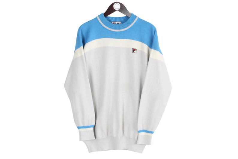 Puede incluir: Un su&eacute;ter Fila vintage en azul claro, blanco y gris, colgado de una percha de madera. El su&eacute;ter presenta un dise&ntilde;o de bloques de color con el logotipo de Fila en el pecho. Los pu&ntilde;os y el cuello tienen rayas azules y blancas.