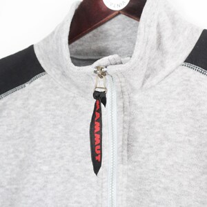 Pu&ograve; includere: Felpa grigia con zip e pannelli neri sulle spalle. La felpa ha un collo alto e una chiusura con zip. Il tirazip ha la scritta "MAMMUT" in rosso. Cuciture bianche visibili.