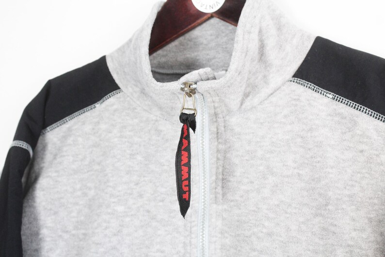 Pu&ograve; includere: Felpa grigia con zip e pannelli neri sulle spalle. La felpa ha un collo alto e una chiusura con zip. Il tirazip ha la scritta "MAMMUT" in rosso. Cuciture bianche visibili.