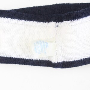 May include: White knitted headband with navy blue trim on the top and bottom edges. A small tag is attached to the headband. The headband is made of a soft, textured material.