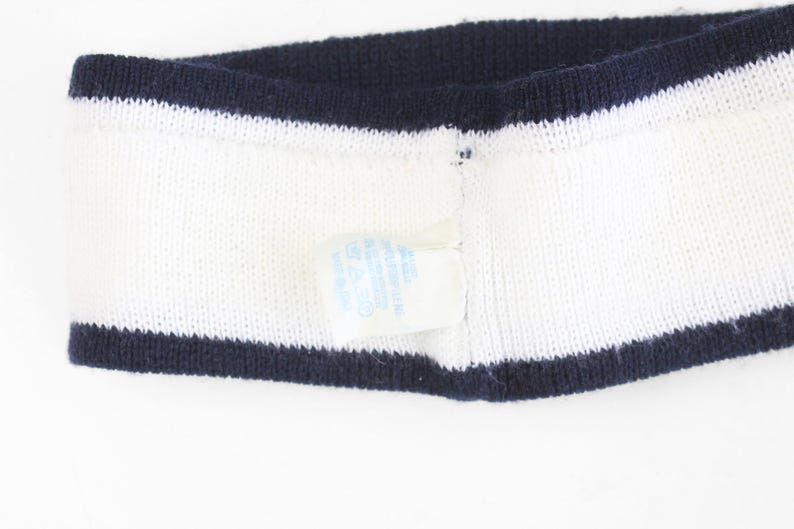 May include: White knitted headband with navy blue trim on the top and bottom edges. A small tag is attached to the headband. The headband is made of a soft, textured material.