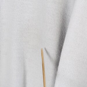 Puede incluir: Primer plano de una tela de punto gris claro con una textura visible. La tela tiene un ligero brillo y parece ser un su&eacute;ter o una prenda similar. Un palillo de madera est&aacute; en el centro inferior de la imagen.