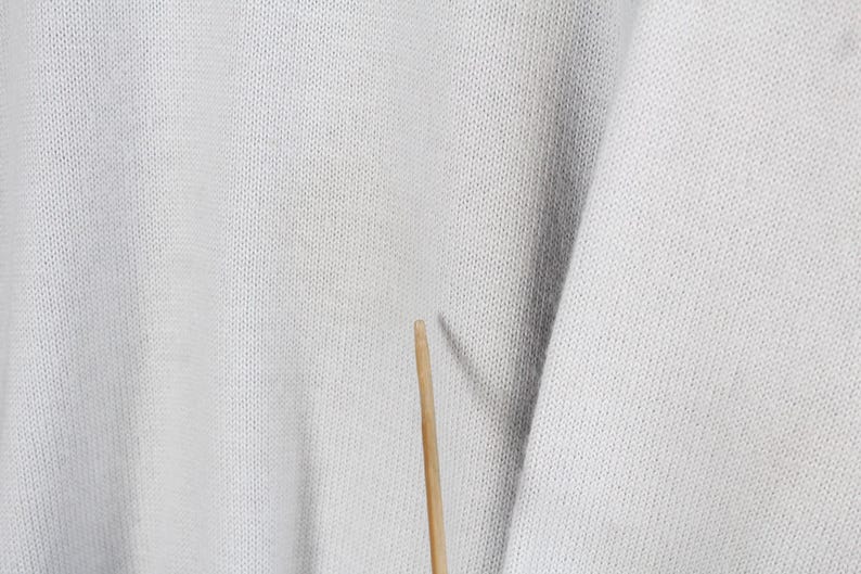 Puede incluir: Primer plano de una tela de punto gris claro con una textura visible. La tela tiene un ligero brillo y parece ser un su&eacute;ter o una prenda similar. Un palillo de madera est&aacute; en el centro inferior de la imagen.