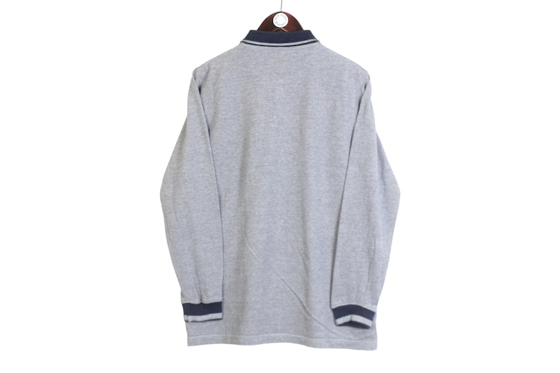May include: A gray long-sleeved shirt with a navy blue collar and cuffs. The shirt is hanging on a wooden hanger against a white background. The cuffs and collar have a navy blue trim.