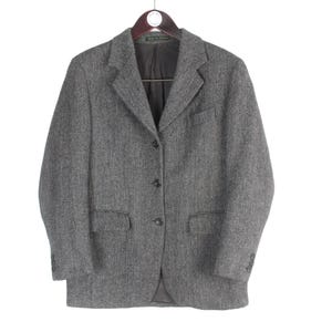Pode incluir: Blazer cinzento de tweed com fecho de três botões, dois bolsos frontais e lapela entalhada. O casaco está pendurado num cabide de madeira sobre um fundo branco. O blazer é de estilo clássico.