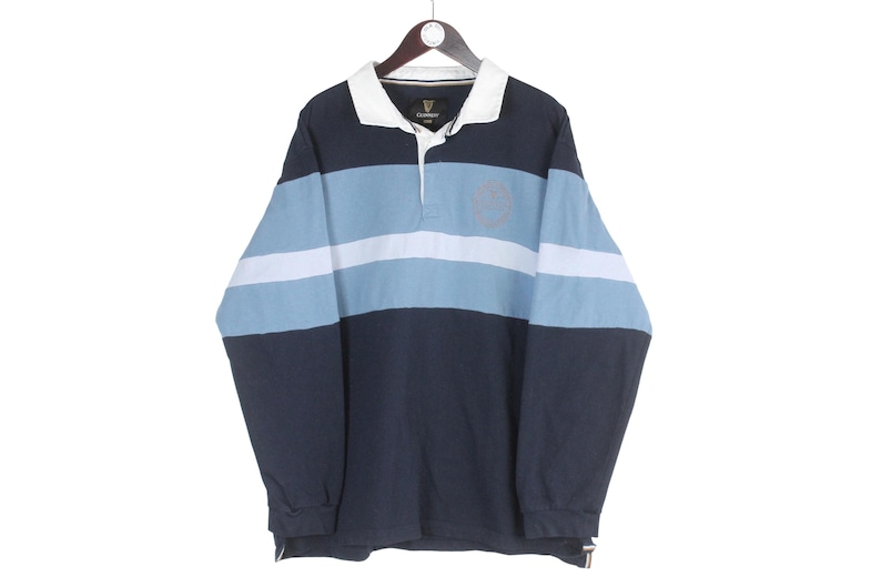 May include: A long-sleeved rugby shirt in navy blue, light blue, and white stripes. The shirt has a white collar and a small logo near the collar. The shirt is hanging on a wooden hanger.