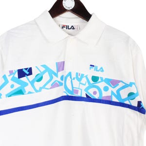 May include: White polo shirt with a colorful geometric design across the chest. The shirt features a collar, short sleeves, and the Fila logo. The design includes blue, teal, and purple shapes.