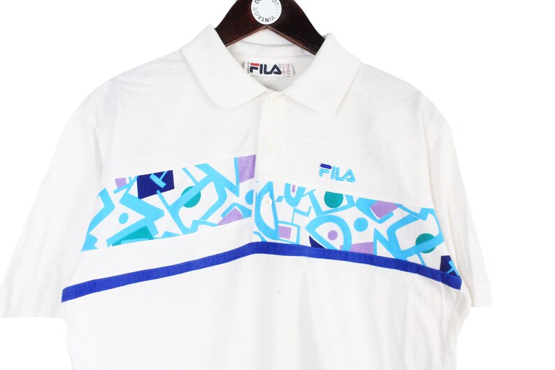 May include: White polo shirt with a colorful geometric design across the chest. The shirt features a collar, short sleeves, and the Fila logo. The design includes blue, teal, and purple shapes.