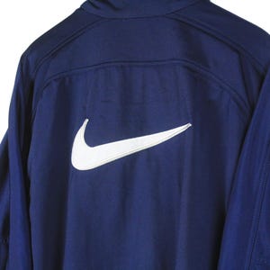 May include: Navy blue athletic jacket with a white Nike logo on the back. The jacket has long sleeves and a high collar. The Nike swoosh is prominently displayed.