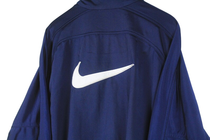 May include: Navy blue athletic jacket with a white Nike logo on the back. The jacket has long sleeves and a high collar. The Nike swoosh is prominently displayed.