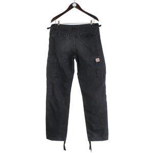 Puede incluir: Pantalones cargo de color gris oscuro colgados de una percha de madera. Los pantalones tienen m&uacute;ltiples bolsillos, un peque&ntilde;o logotipo cuadrado en el bolsillo derecho y cordones en los tobillos. Los pantalones est&aacute;n hechos de una tela duradera.