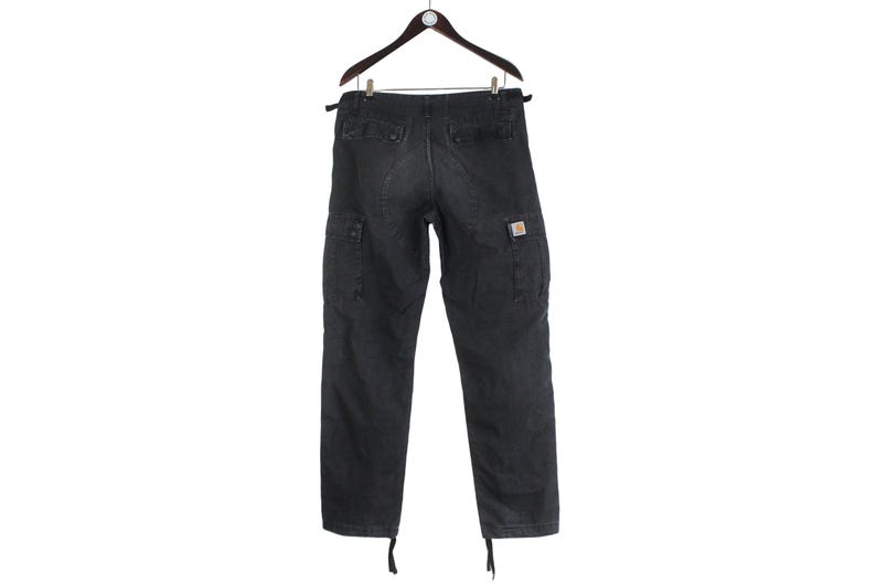 Puede incluir: Pantalones cargo de color gris oscuro colgados de una percha de madera. Los pantalones tienen m&uacute;ltiples bolsillos, un peque&ntilde;o logotipo cuadrado en el bolsillo derecho y cordones en los tobillos. Los pantalones est&aacute;n hechos de una tela duradera.