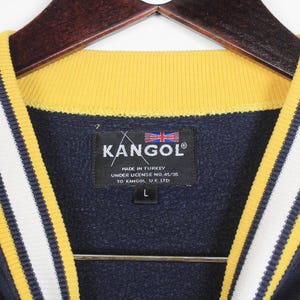 K&ouml;nnte beinhalten: Marineblaues Kangol-Sweatshirt mit gelb-wei&szlig; gestreiften Zierleisten. Der Kragen ist gelb. Das Kangol-Etikett ist sichtbar, mit der Union Jack-Flagge und dem Text "Made in Turkey". Das Sweatshirt ist Gr&ouml;&szlig;e L.