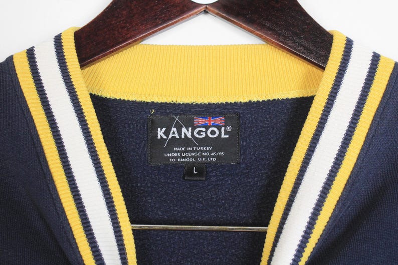 K&ouml;nnte beinhalten: Marineblaues Kangol-Sweatshirt mit gelb-wei&szlig; gestreiften Zierleisten. Der Kragen ist gelb. Das Kangol-Etikett ist sichtbar, mit der Union Jack-Flagge und dem Text "Made in Turkey". Das Sweatshirt ist Gr&ouml;&szlig;e L.