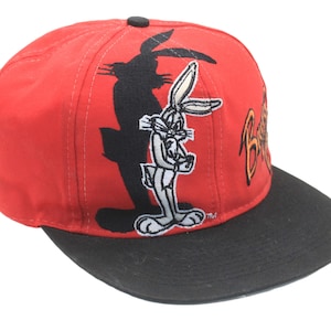 May include: A red and black Bugs Bunny baseball cap. The cap has an embroidered Bugs Bunny character and shadow design on the front. The brim is black, and the word "Bugs" is in orange.