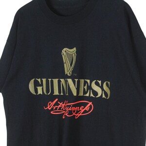 Op de afbeelding: Zwart T-shirt met het gouden Guinness-logo, inclusief een harp en het woord "GUINNESS". De handtekening "Arthur Guinness" staat eronder in rode letters. Het shirt heeft korte mouwen.
