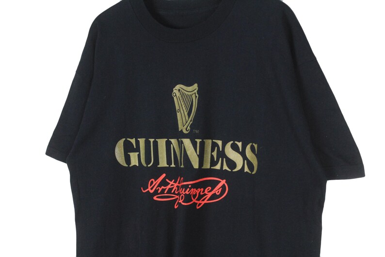 Op de afbeelding: Zwart T-shirt met het gouden Guinness-logo, inclusief een harp en het woord "GUINNESS". De handtekening "Arthur Guinness" staat eronder in rode letters. Het shirt heeft korte mouwen.