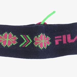 May include: A navy blue knit headband with pink and green floral and arrow patterns. The word "FILA" is printed in pink. A green string is attached to the top of the headband.