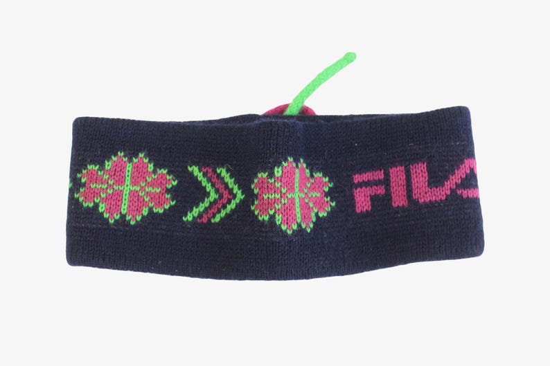 May include: A navy blue knit headband with pink and green floral and arrow patterns. The word "FILA" is printed in pink. A green string is attached to the top of the headband.