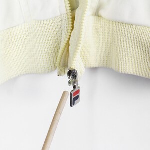 May include: A white jacket featuring a cream-colored ribbed hem and a silver metal zipper pull. The zipper pull is rectangular and displays the Fila logo in red, white, and navy blue. A wooden pointer is directed toward the zipper pull.