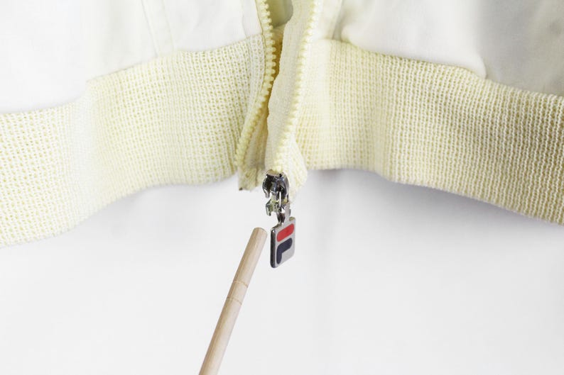 May include: A white jacket featuring a cream-colored ribbed hem and a silver metal zipper pull. The zipper pull is rectangular and displays the Fila logo in red, white, and navy blue. A wooden pointer is directed toward the zipper pull.