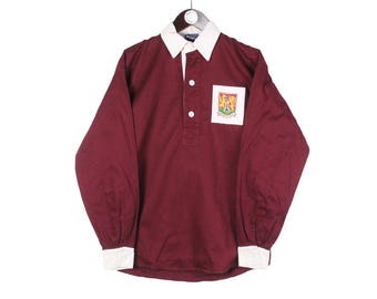 May include: Maroon long-sleeved shirt with a white collar and cuffs. The shirt has a small pocket with a crest on the left chest. The shirt is hanging on a metal hanger.