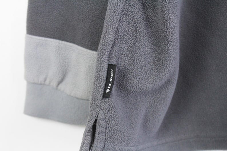 Puede incluir: Primer plano de una sudadera de forro polar gris con un dise&ntilde;o de bloques de color. La sudadera presenta un cuerpo gris oscuro, una banda gris claro y una manga gris. Una etiqueta negra con el logotipo "Guinness" est&aacute; cosida en la manga.