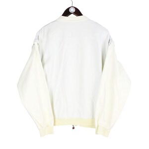 May include: A cream-colored long-sleeve jacket featuring a solid white back panel, a ribbed collar, and ribbed cuffs. The garment is displayed on a dark brown hanger and includes a small circular tag attached to the collar area.