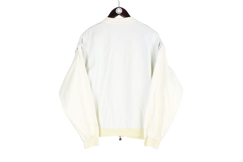 May include: A cream-colored long-sleeve jacket featuring a solid white back panel, a ribbed collar, and ribbed cuffs. The garment is displayed on a dark brown hanger and includes a small circular tag attached to the collar area.