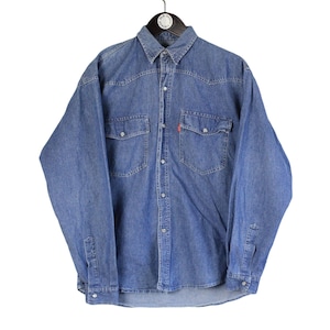 May include: A long-sleeve blue denim button-down shirt featuring a pointed collar, two chest pockets with buttoned flaps, and a small red tab detail on the left pocket. The shirt has a front button closure and long sleeves with buttoned cuffs.