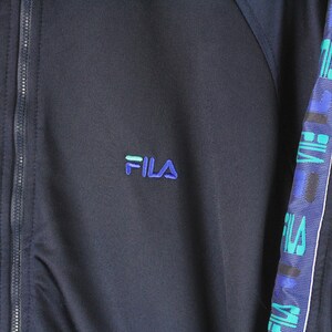 Puede incluir: Una chaqueta azul marino con cremallera, con una cremallera blanca y un logotipo FILA azul y verde en el pecho izquierdo. La chaqueta tiene una franja blanca en la manga derecha con texto FILA azul y verde.