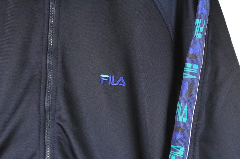 Puede incluir: Una chaqueta azul marino con cremallera, con una cremallera blanca y un logotipo FILA azul y verde en el pecho izquierdo. La chaqueta tiene una franja blanca en la manga derecha con texto FILA azul y verde.