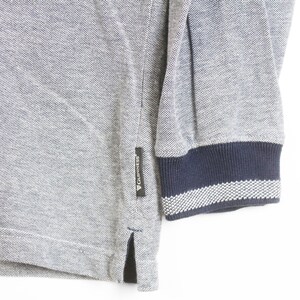 May include: Close-up of a gray long-sleeved shirt with a navy blue and white striped cuff. The shirt has a small black tag with white text. The fabric has a textured appearance.