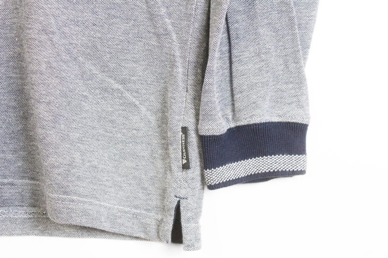 May include: Close-up of a gray long-sleeved shirt with a navy blue and white striped cuff. The shirt has a small black tag with white text. The fabric has a textured appearance.