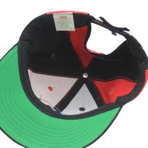 May include: Black, red, and green baseball cap with a flat brim. The cap features a red and black design on the crown and a green brim. The interior shows a white and red pattern. The Nike logo is visible.