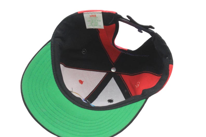 May include: Black, red, and green baseball cap with a flat brim. The cap features a red and black design on the crown and a green brim. The interior shows a white and red pattern. The Nike logo is visible.