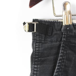 Puede incluir: Primer plano de pantalones cargo negros con un patr&oacute;n de cuadr&iacute;cula, colgados de una percha de madera. Los pantalones tienen una trabilla negra con una hebilla plateada. La tela parece desgastada.