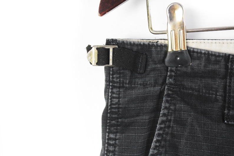 Puede incluir: Primer plano de pantalones cargo negros con un patr&oacute;n de cuadr&iacute;cula, colgados de una percha de madera. Los pantalones tienen una trabilla negra con una hebilla plateada. La tela parece desgastada.