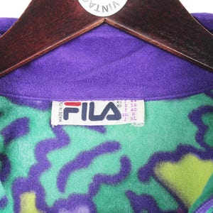 Puede incluir: Una chaqueta de forro polar morada con un vibrante patr&oacute;n abstracto en verde y morado. La etiqueta interior del cuello muestra el logotipo de la marca FILA en rojo y negro, con el texto Made in Italy e informaci&oacute;n de talla en la etiqueta.