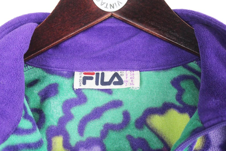 Puede incluir: Una chaqueta de forro polar morada con un vibrante patr&oacute;n abstracto en verde y morado. La etiqueta interior del cuello muestra el logotipo de la marca FILA en rojo y negro, con el texto Made in Italy e informaci&oacute;n de talla en la etiqueta.