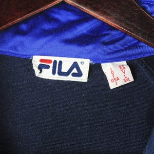 Puede incluir: Una chaqueta Fila azul marino con cuello azul y una etiqueta blanca con el logotipo de Fila e informaci&oacute;n de talla. La etiqueta dice "Fila", "IDF", "USA J", "33", "54", "V", "L", "3XL".