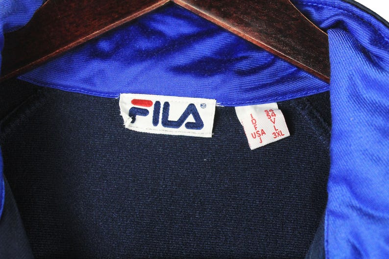 Puede incluir: Una chaqueta Fila azul marino con cuello azul y una etiqueta blanca con el logotipo de Fila e informaci&oacute;n de talla. La etiqueta dice "Fila", "IDF", "USA J", "33", "54", "V", "L", "3XL".