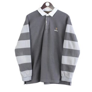 Puede incluir: Una camiseta de rugby gris con mangas largas a rayas blancas y grises. Tiene cuello blanco y un peque&ntilde;o logo en el pecho. La camiseta est&aacute; colgada de una percha de madera.